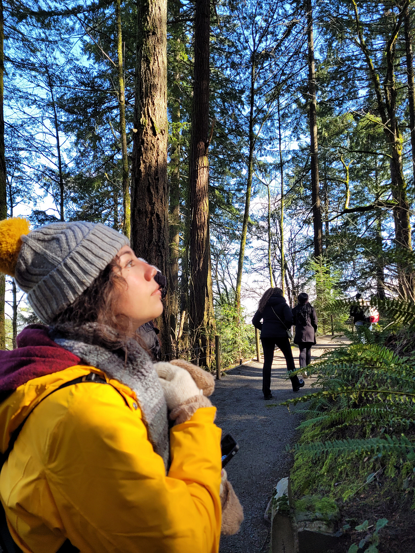 (Seattle, 2022) Andrea, dressed in a yellow winter coat, stops in the forest trail to look toward the lazy sun.