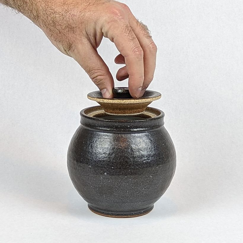 Round lidded jar in matte black. Hand showing sclale, as well as function of the knob of the lid.