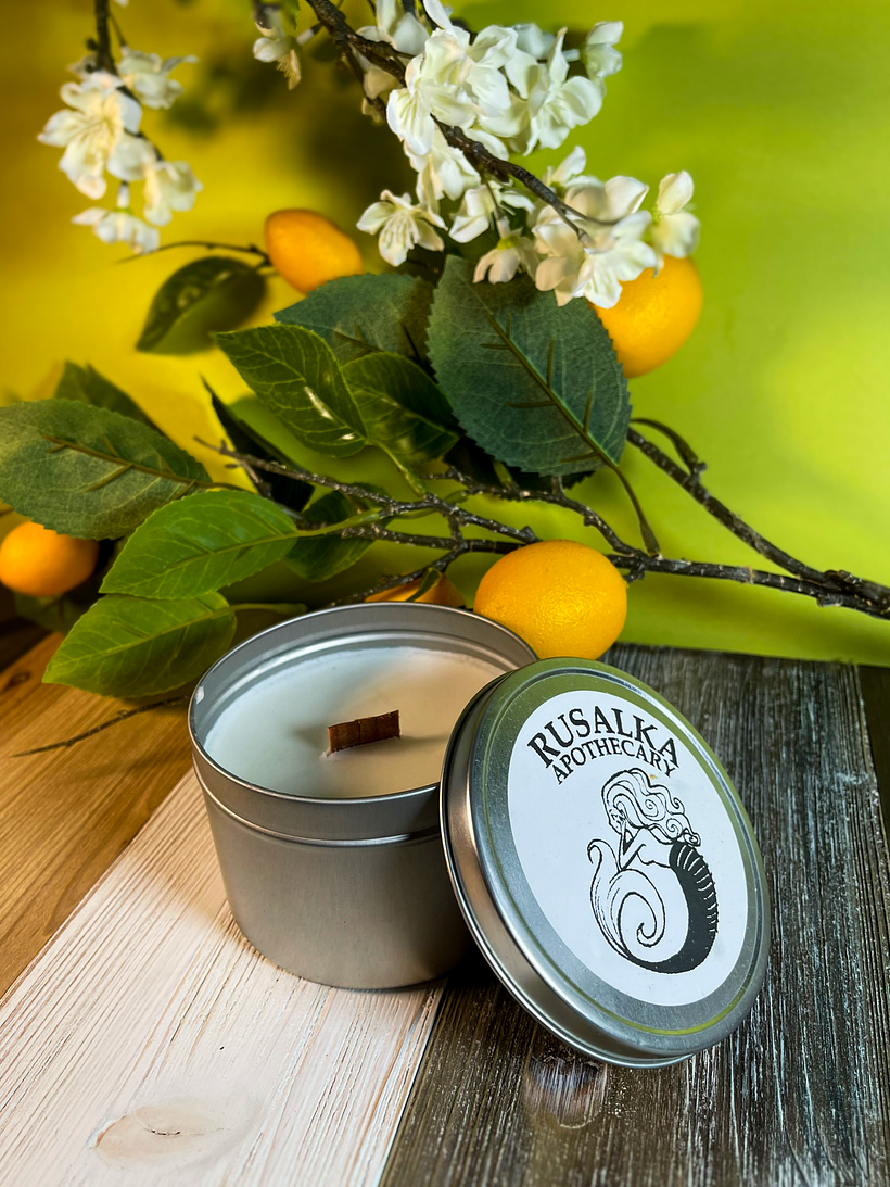 Wax candle in silver tin and label reading Rusalka Apothecary shown on wood table against lemon branches with fruit